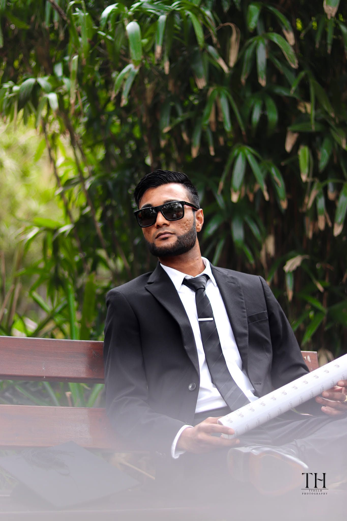 Stylish man in black suit and sunglasses sitting on a bench reading a newspaper outdoors.