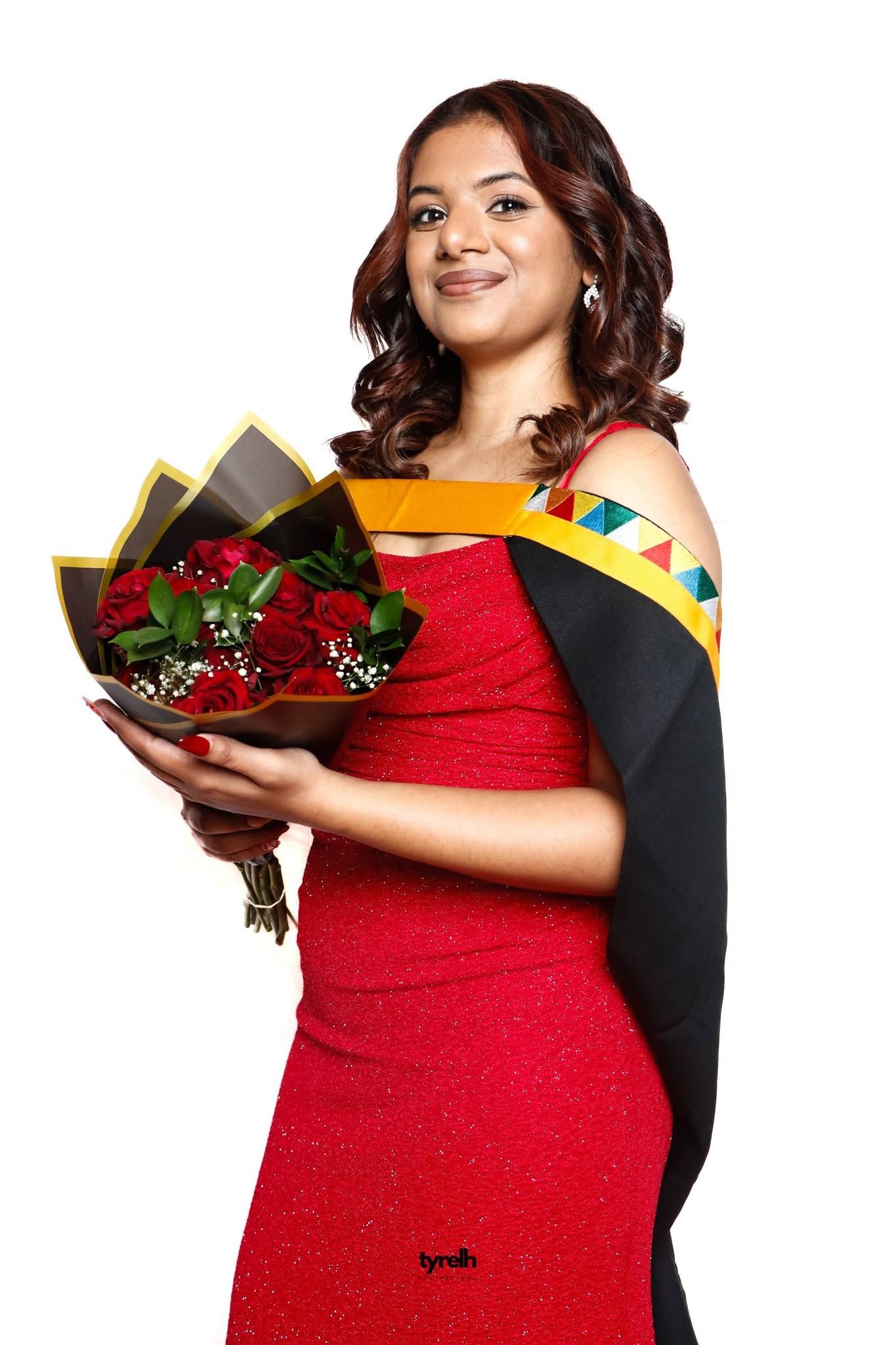 Woman in red dress and graduation sash smiles holding bouquet of red roses on white background.