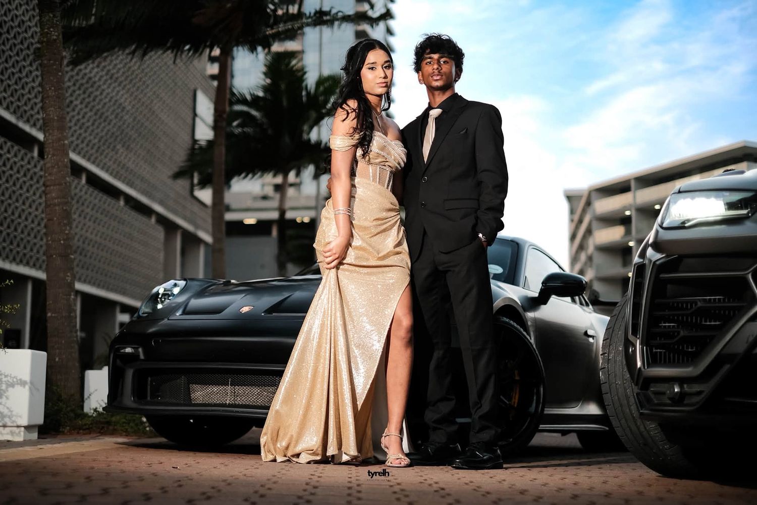 Elegant couple in formal wear posing by luxury black sports cars for prom photos.