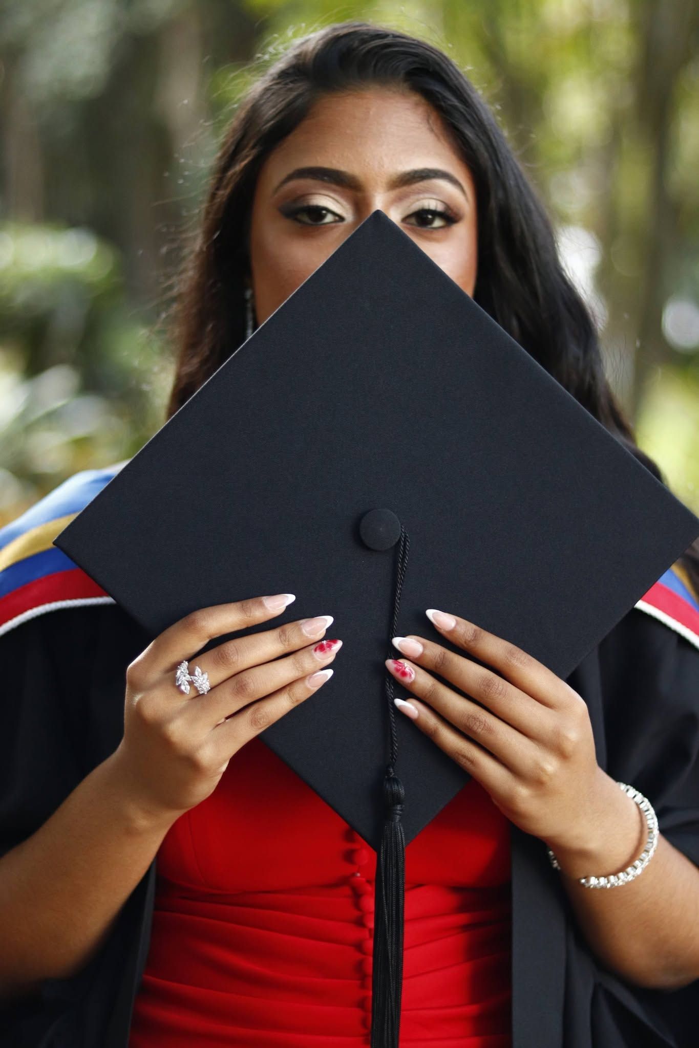 A young woman in graduation gown holds her mortarboard cap in front of her face, showing only her eyes.