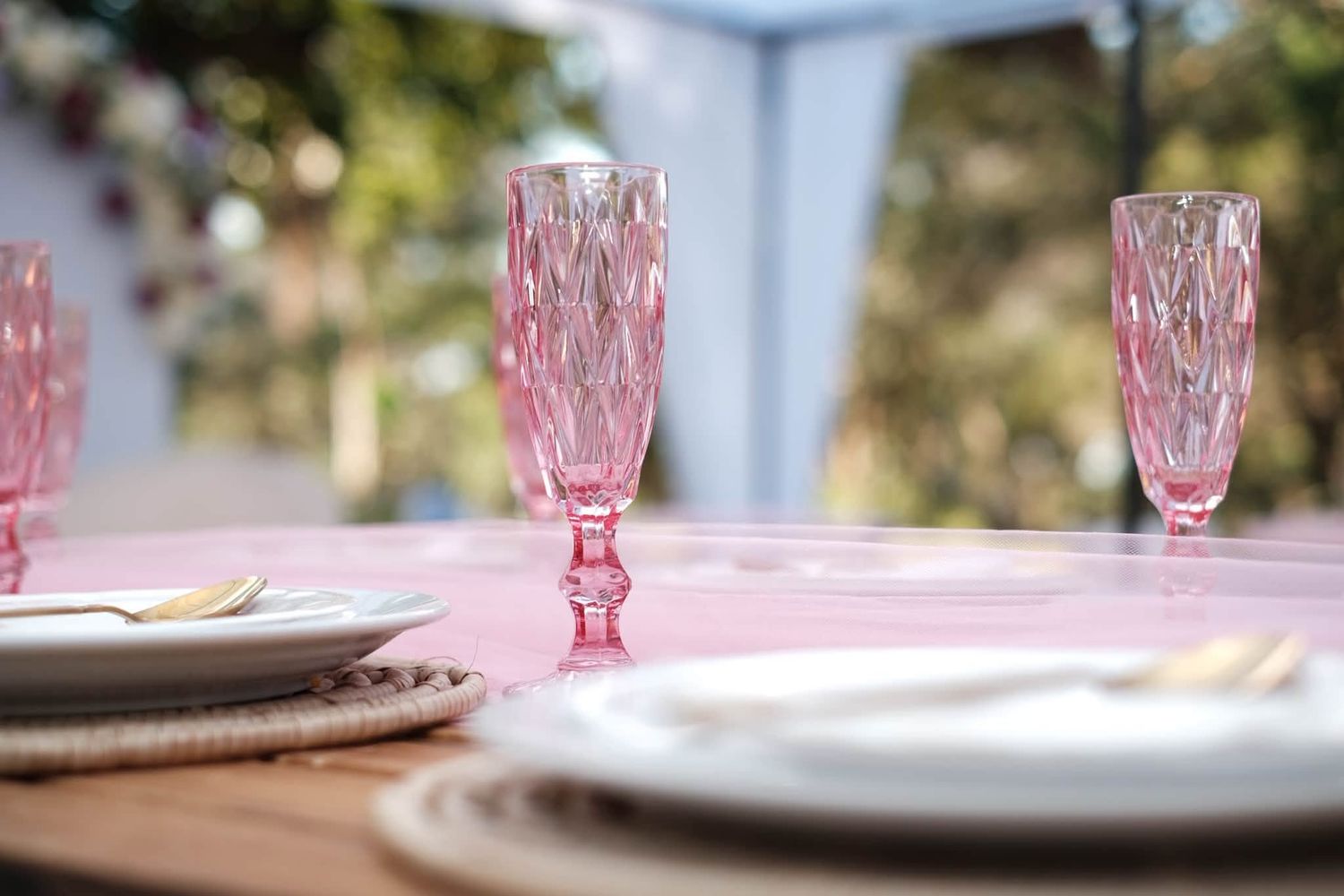 Elegant pink crystal champagne flutes on a pink tablecloth with white plates and gold cutlery at an outdoor table setting.