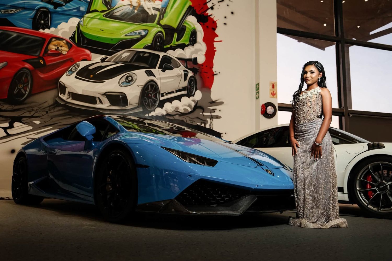 Woman in silver gown posing beside a blue Lamborghini Huracan at a luxury car showroom with colorful mural.