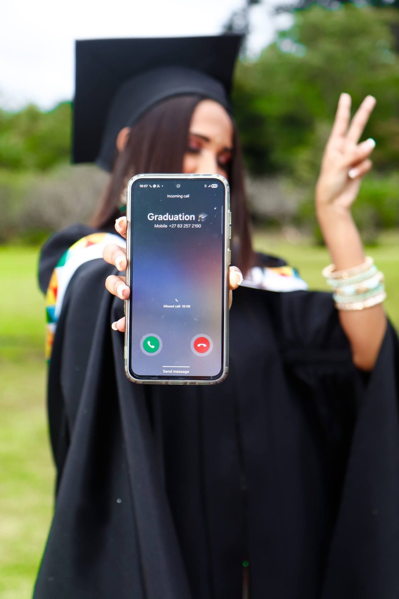 Graduate in cap and gown holds phone showing incoming call labeled Graduation while making peace sign outdoors.