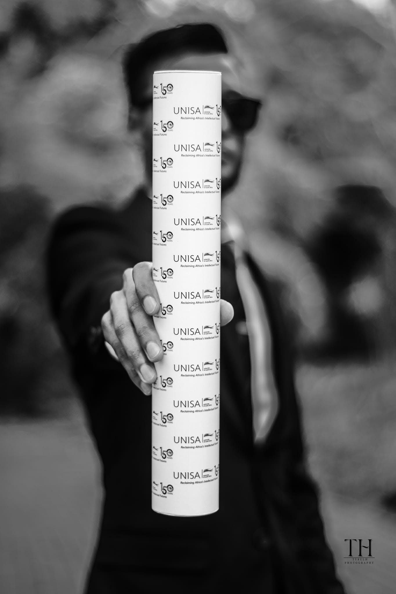 Black and white photo of a graduate holding a stack of UNISA degree certificates outdoors.