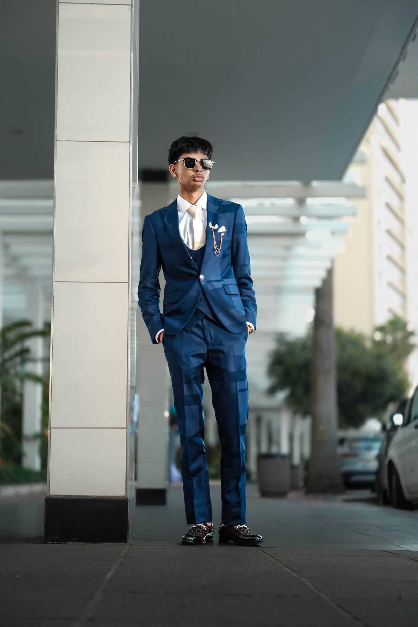 Young man in navy blue suit with white shirt and sunglasses posing outdoors near a column.