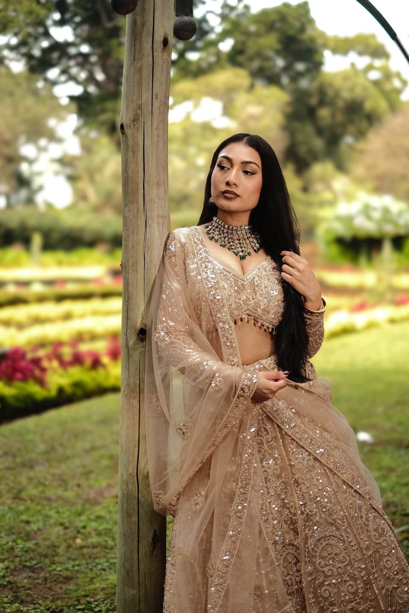 Woman in elegant beige embroidered lehenga with dupatta poses in a lush garden setting.