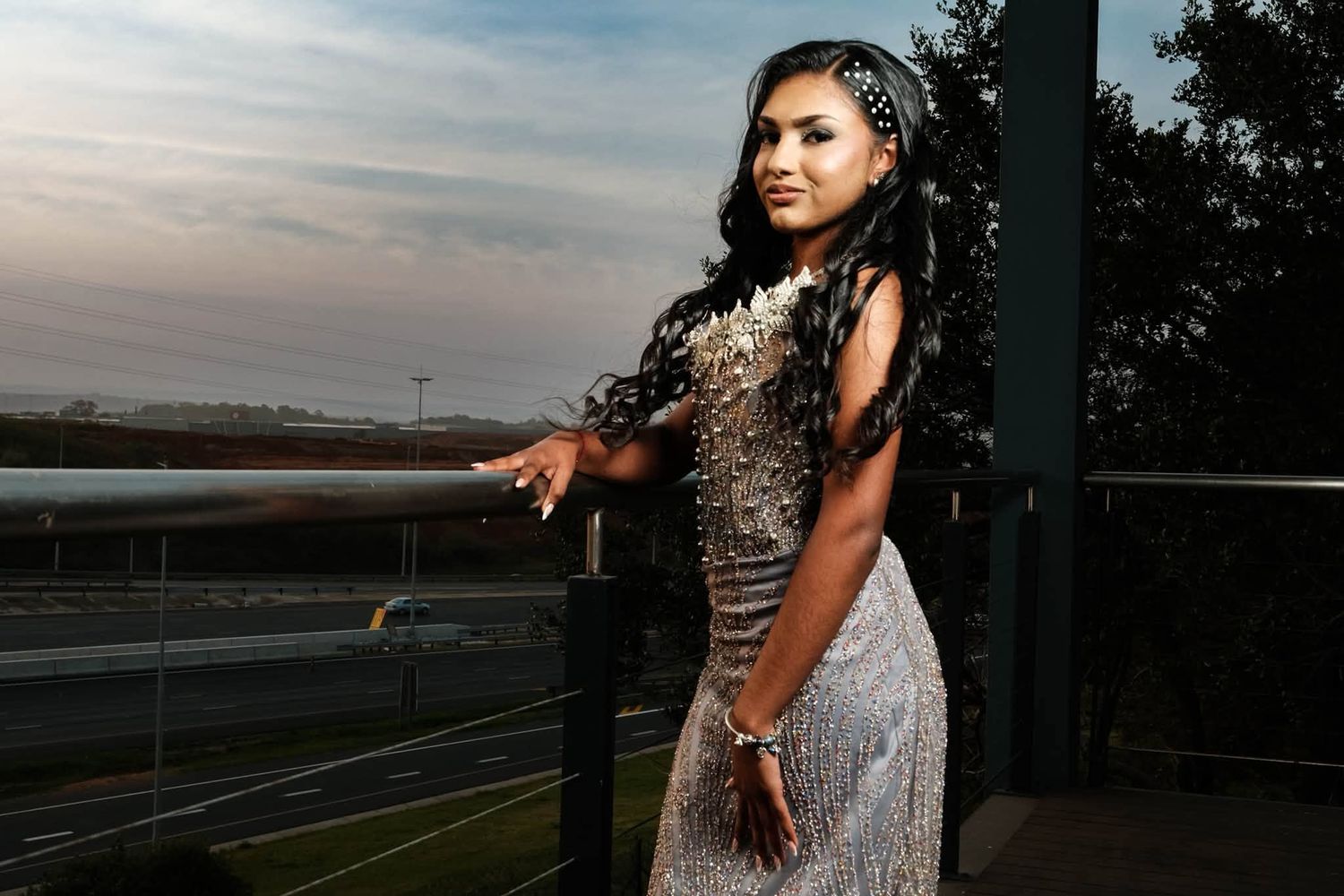 Young woman in a beaded silver gown poses on a balcony at dusk with long braided hair.