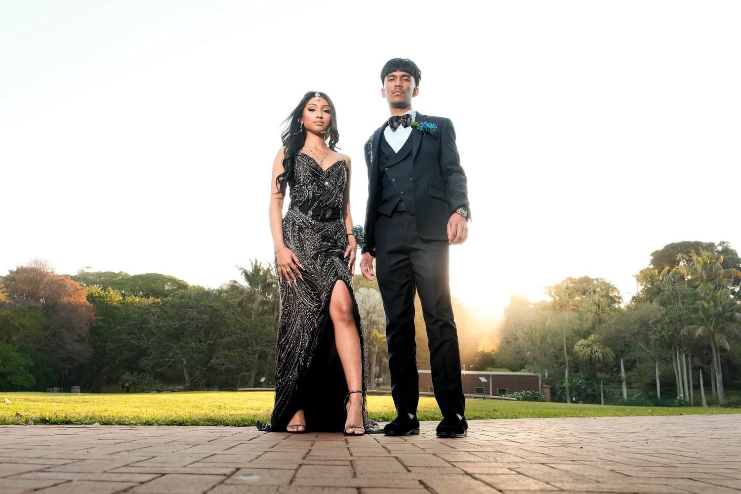 Elegant couple in formal black attire posing outdoors at golden hour for a prom or gala event.