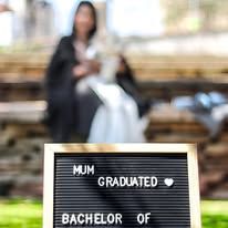A letterboard sign reading MUM GRADUATED BACHELOR OF with a graduate in cap and gown sitting in background.