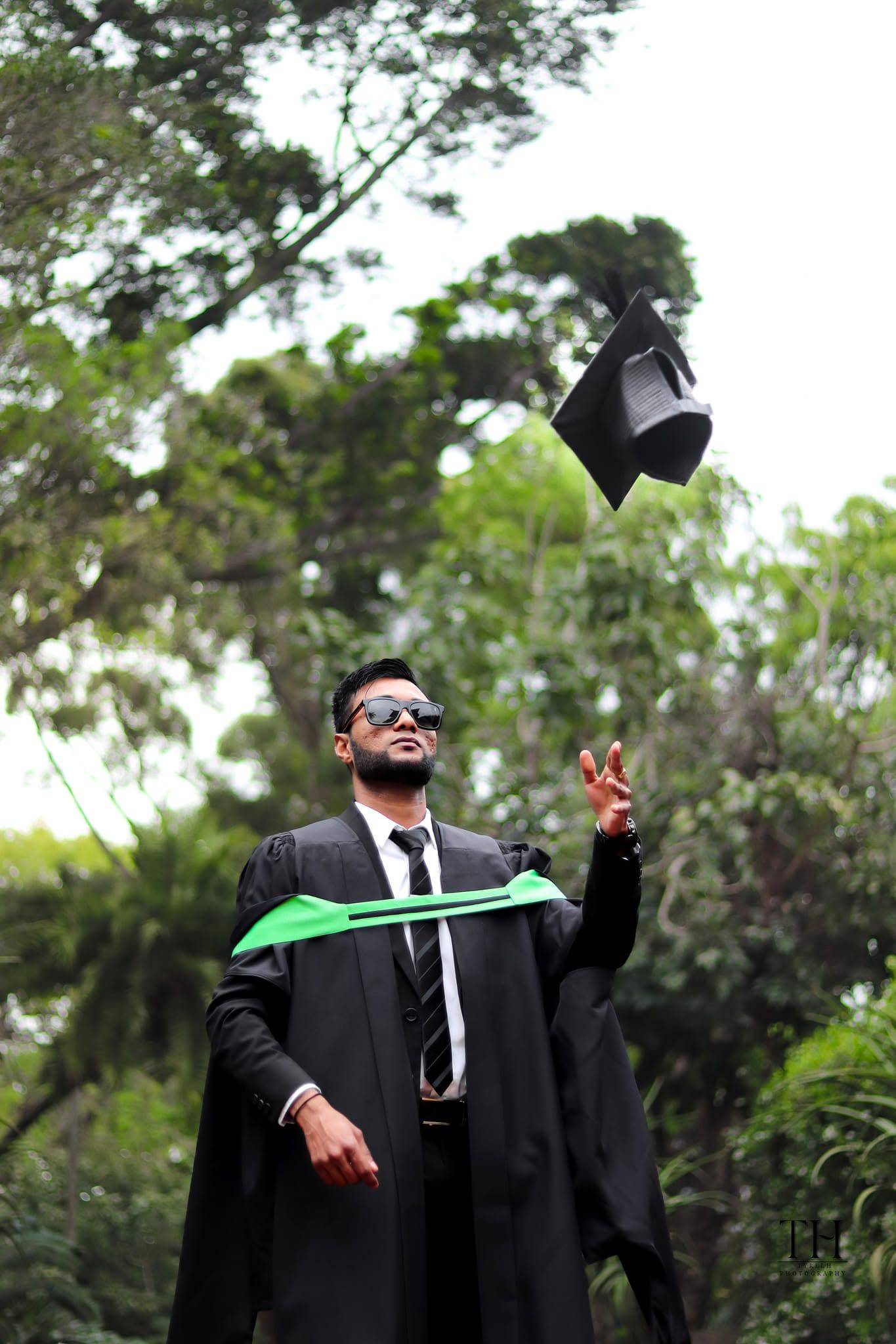 Graduate in black gown tosses mortarboard cap in the air outdoors, celebrating academic achievement.
