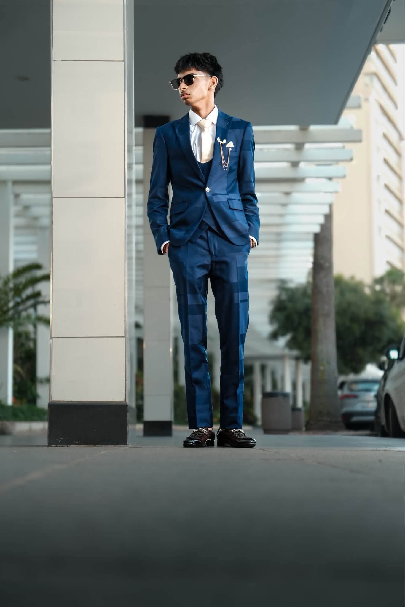 Stylish man in navy plaid suit with pocket square and sunglasses standing outdoors.