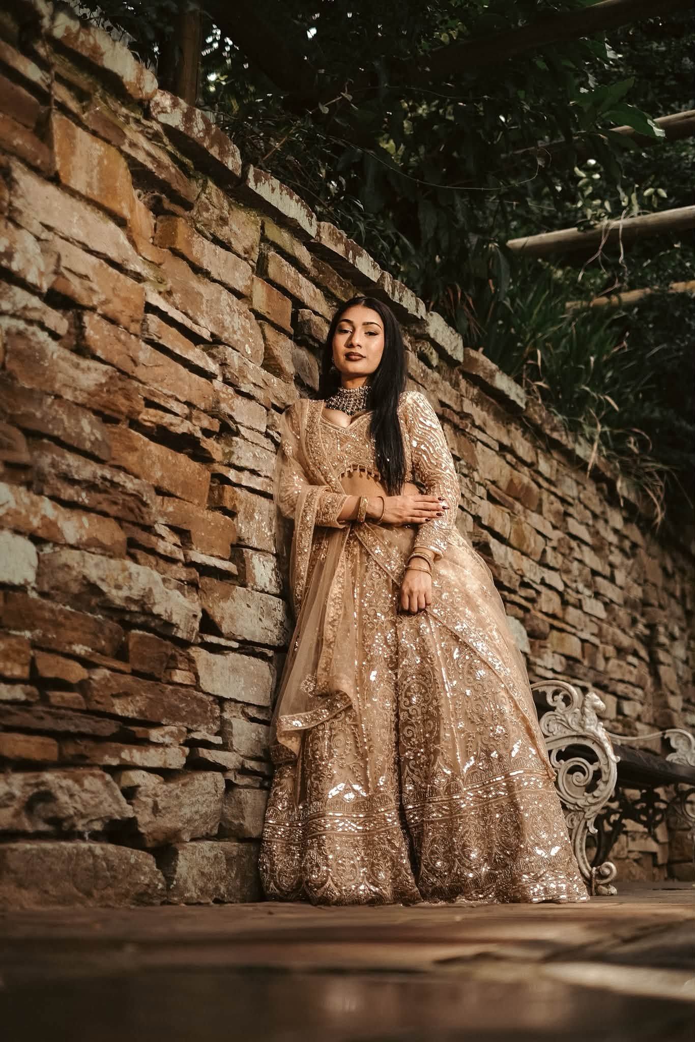 Woman in golden embroidered lehenga posing against rustic stone wall surrounded by lush greenery.