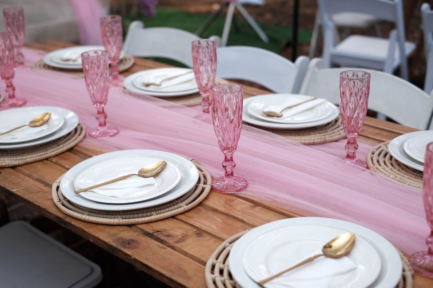 Elegant pink table setting with gold cutlery, pink crystal glasses, and white plates on a rustic wooden farm table.