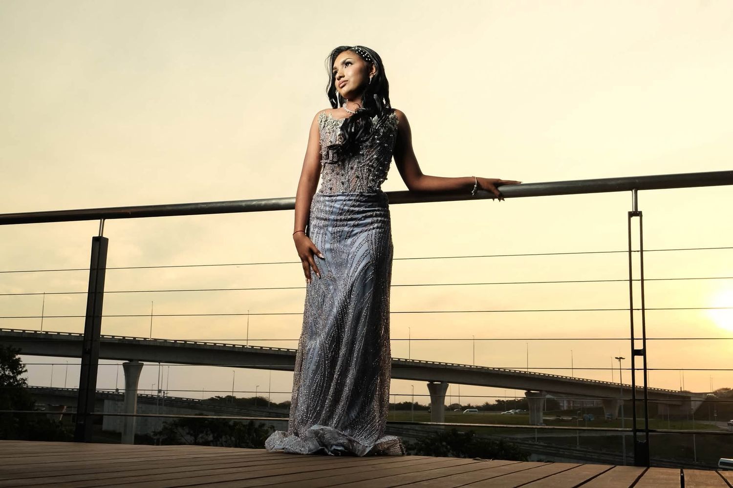 Woman in elegant silver gown poses on deck at sunset with bridge in background.