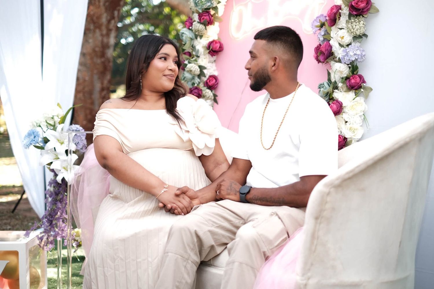 Pregnant couple sitting together at a floral baby shower, holding hands and smiling at each other in matching white outfits.