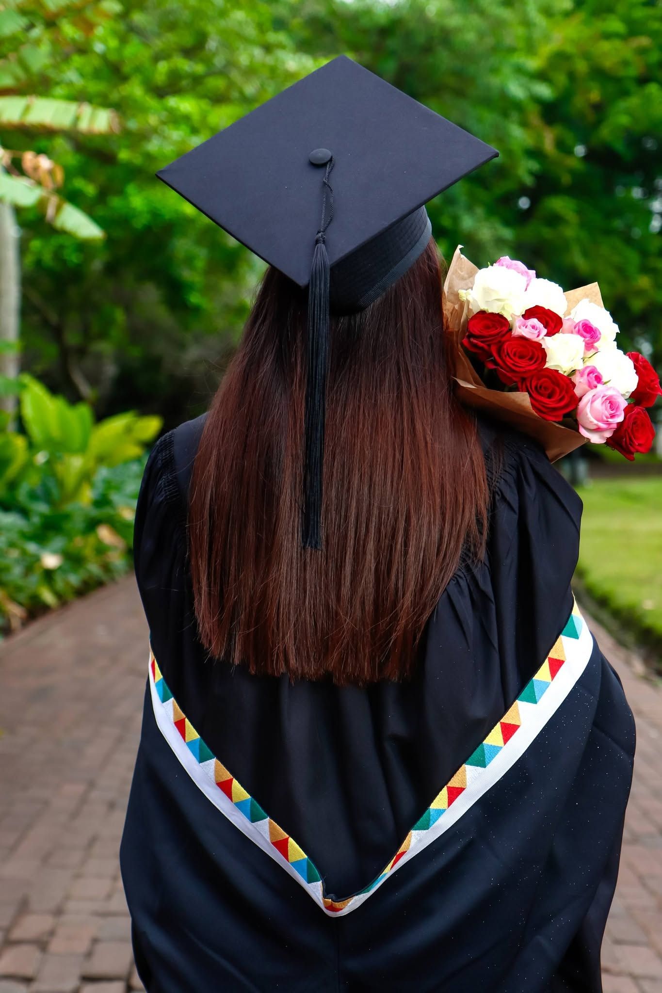 Graduate in black cap and gown holding colorful rose bouquet, viewed from behind in a lush green garden setting.