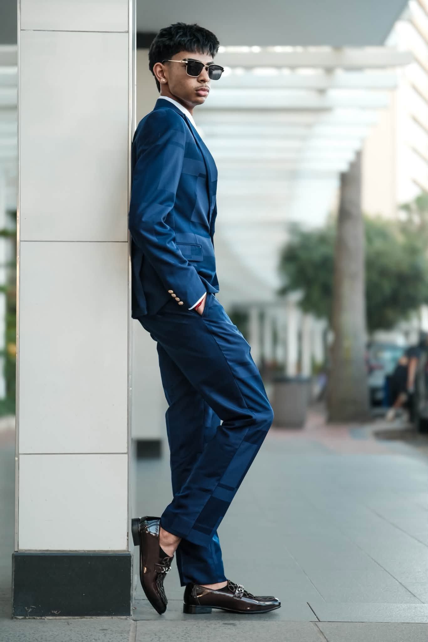Young man in navy blue suit and sunglasses leaning against white pillar outdoors.