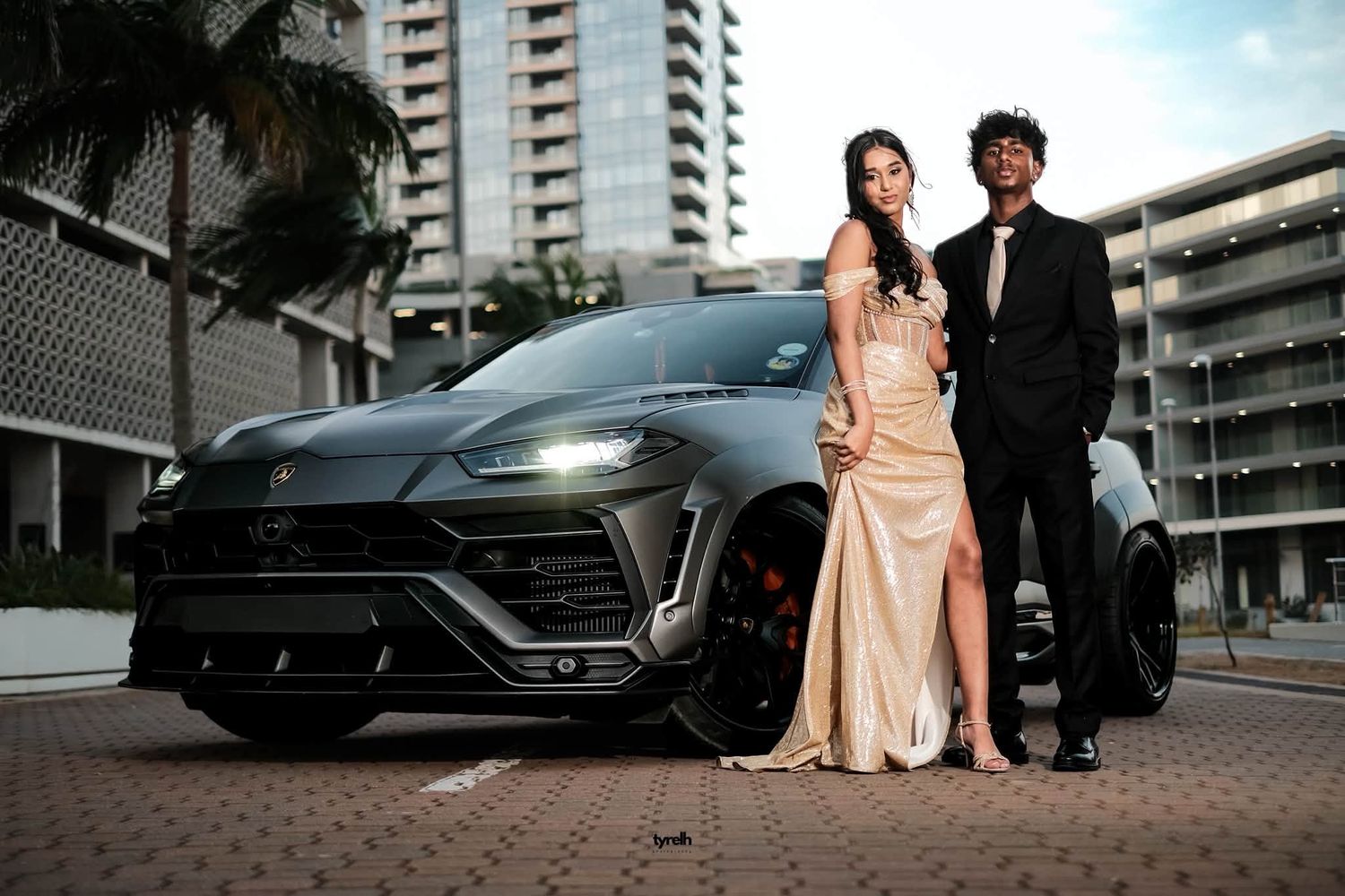 Couple in formal attire posing in front of a black Lamborghini Urus SUV with city buildings in background.