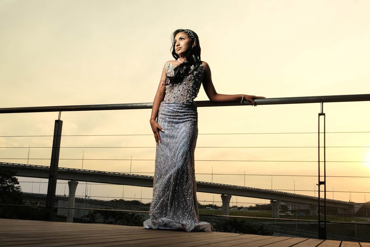 Woman in elegant silver gown posing on outdoor deck at sunset with bridge in background.