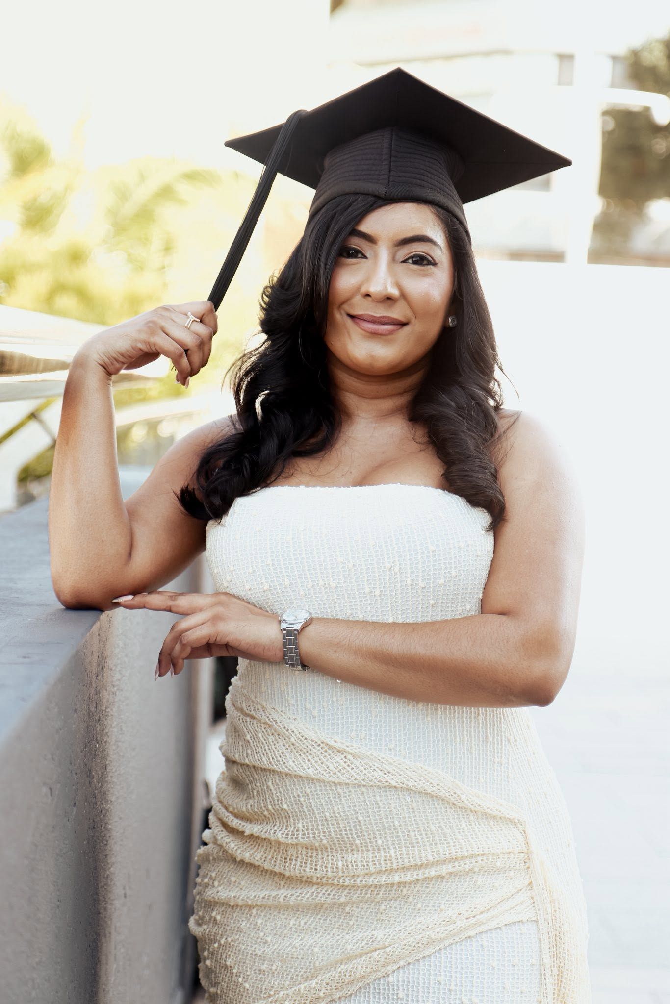 Young woman in white sequin dress and graduation cap smiling outdoors, celebrating academic achievement.