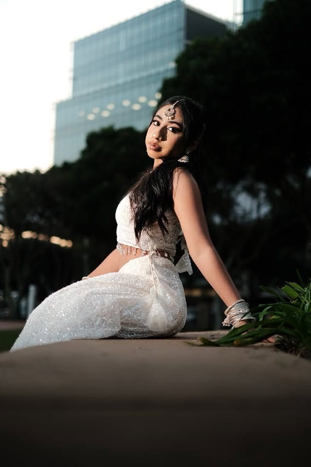 A young woman in a white sparkly Indian outfit with traditional jewelry poses outdoors near a modern building.