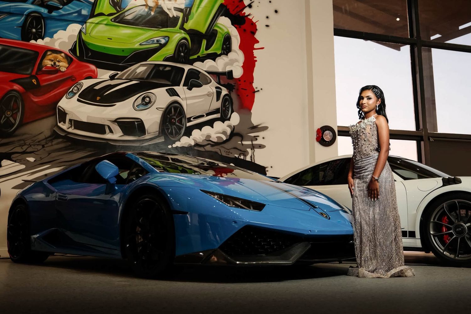 Woman in elegant gown poses beside a blue Lamborghini Huracan at a luxury car showroom with colorful mural backdrop.