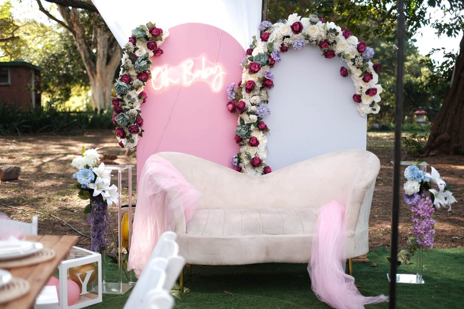 Elegant baby shower setup with floral arch, cream sofa, pink tulle, and Oh Baby neon sign outdoors.