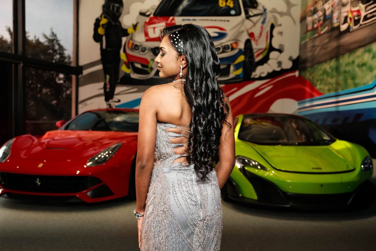 Woman in silver dress poses between red Ferrari and green Lamborghini in front of racing car artwork.