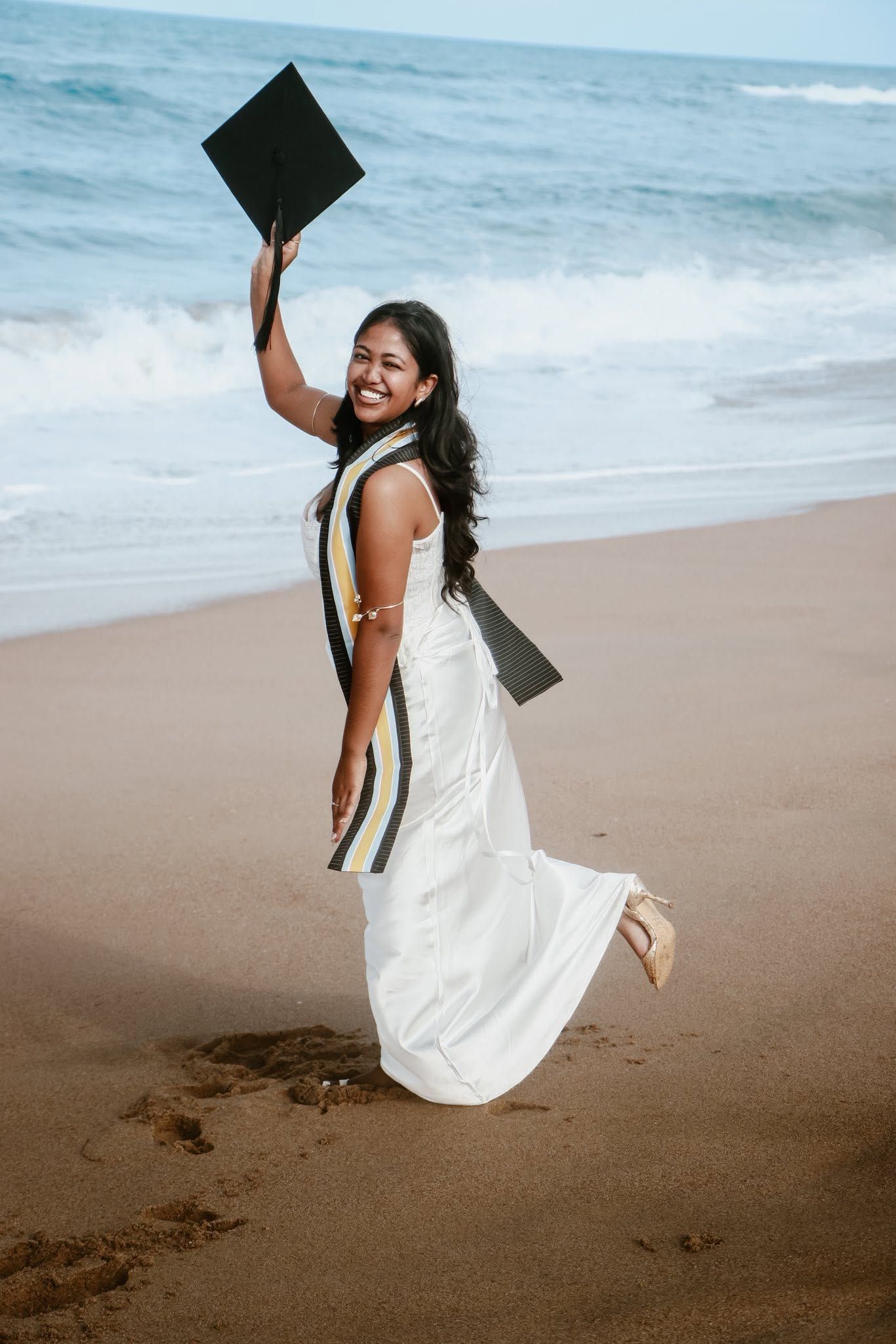 Smiling graduate in white dress holding cap on beach, celebrating with academic sash, ocean waves in background.
