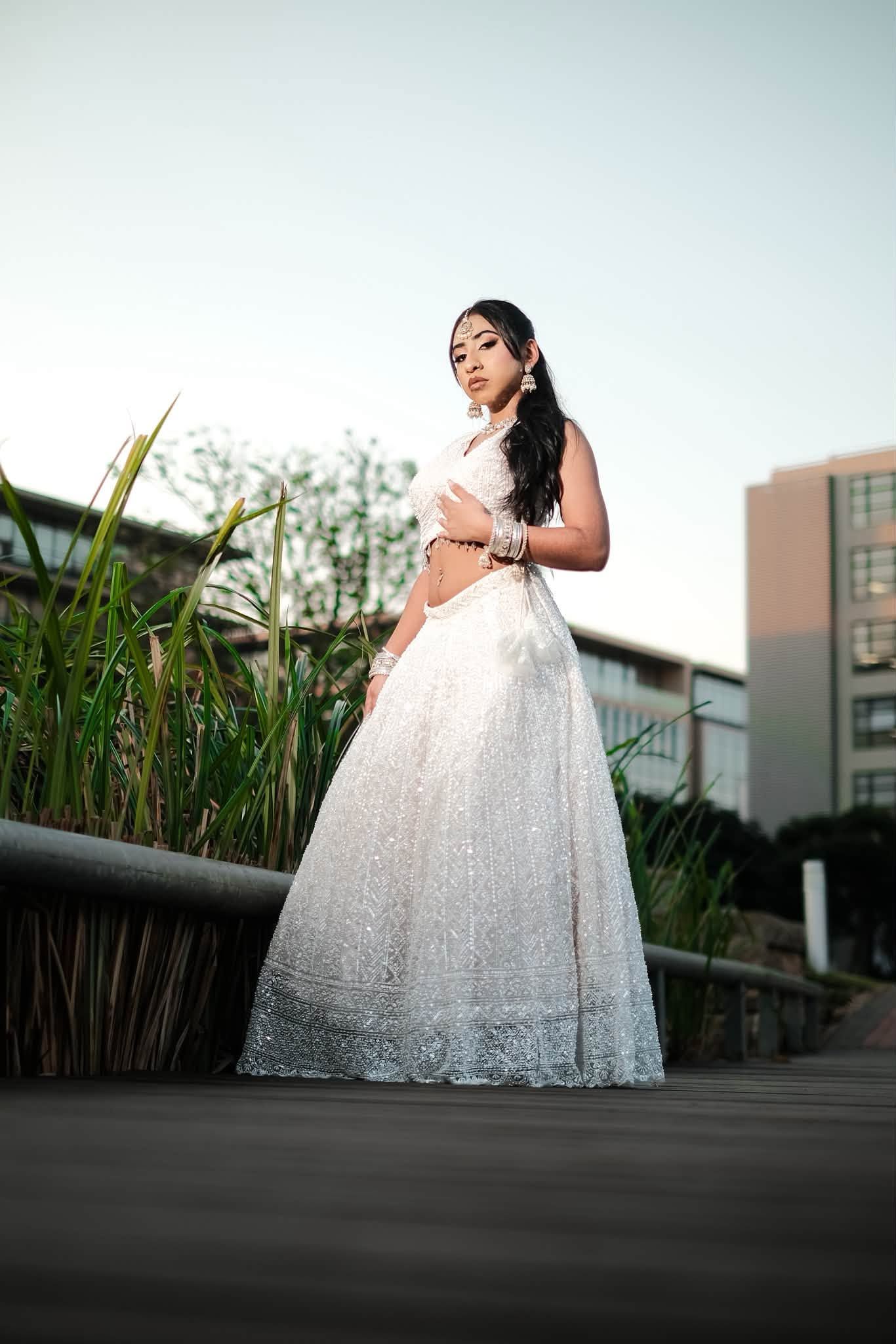 Young woman in elegant white embroidered lehenga poses outdoors on wooden deck with lush greenery and building backdrop.