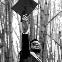 Graduate in cap and gown tosses mortarboard into the air among birch trees in black and white photo.