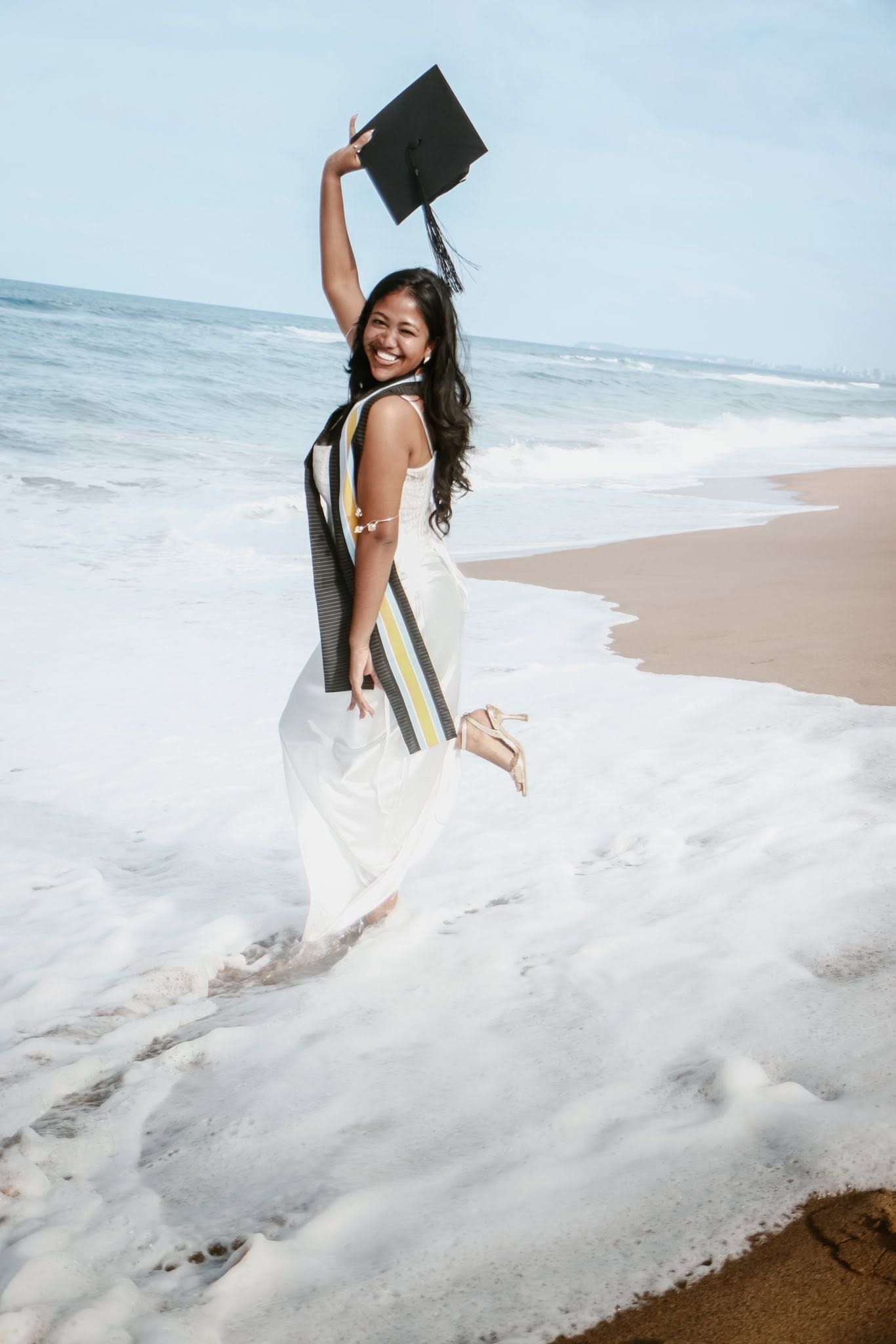 Joyful graduate in white dress holding mortarboard on beach, waves crashing around her feet.