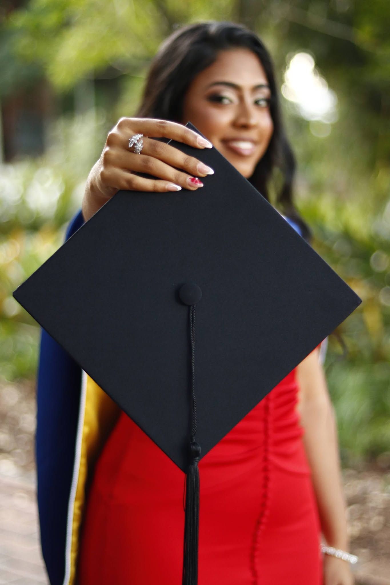 Smiling graduate in red dress holding black mortarboard cap toward camera with green trees in background.
