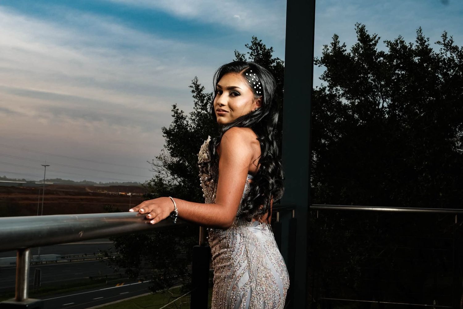 Young woman in elegant beaded gown poses at railing during sunset with trees in background.