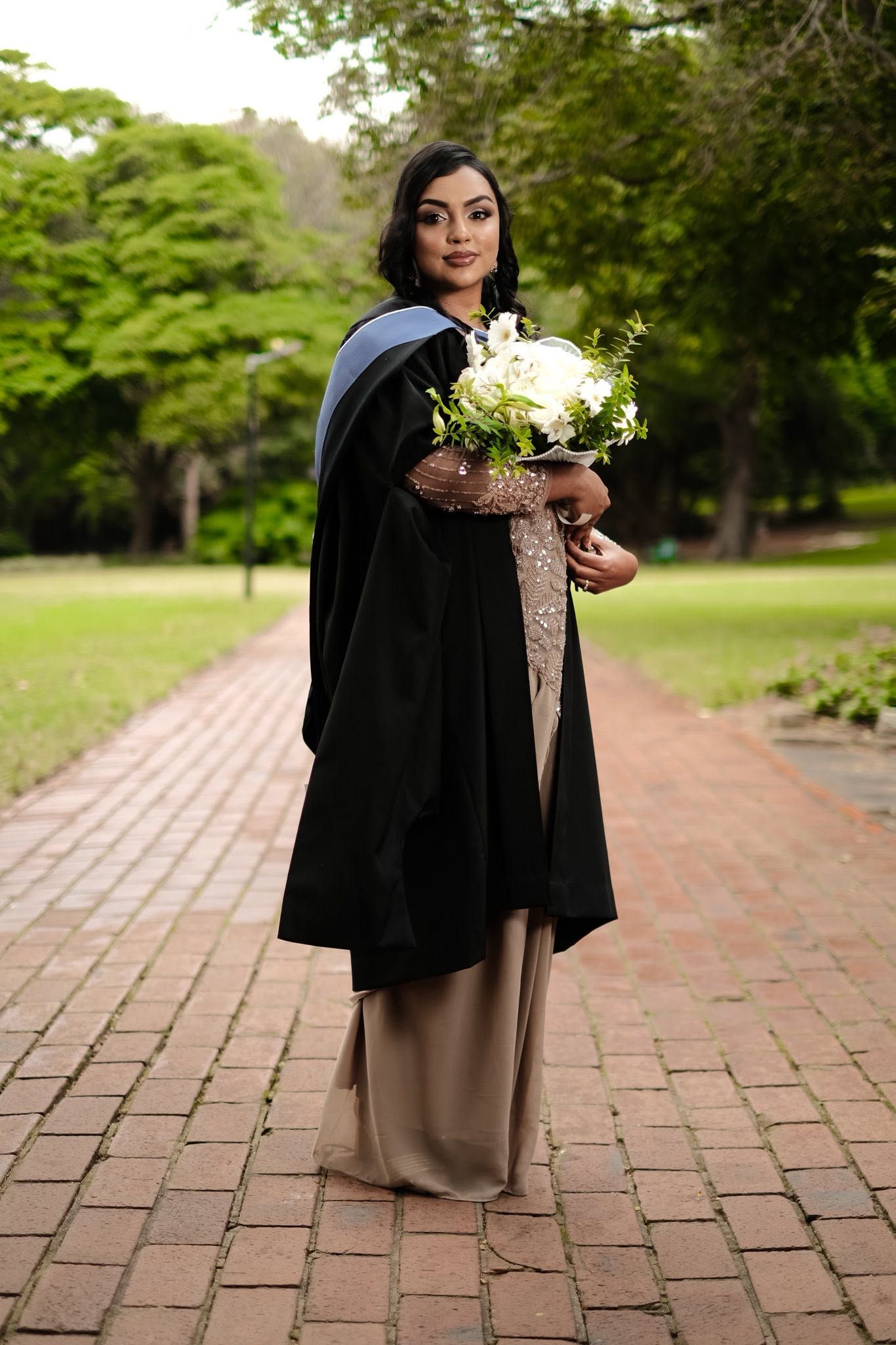 A young woman in a black graduation gown holds white flowers on a brick path surrounded by lush green trees.