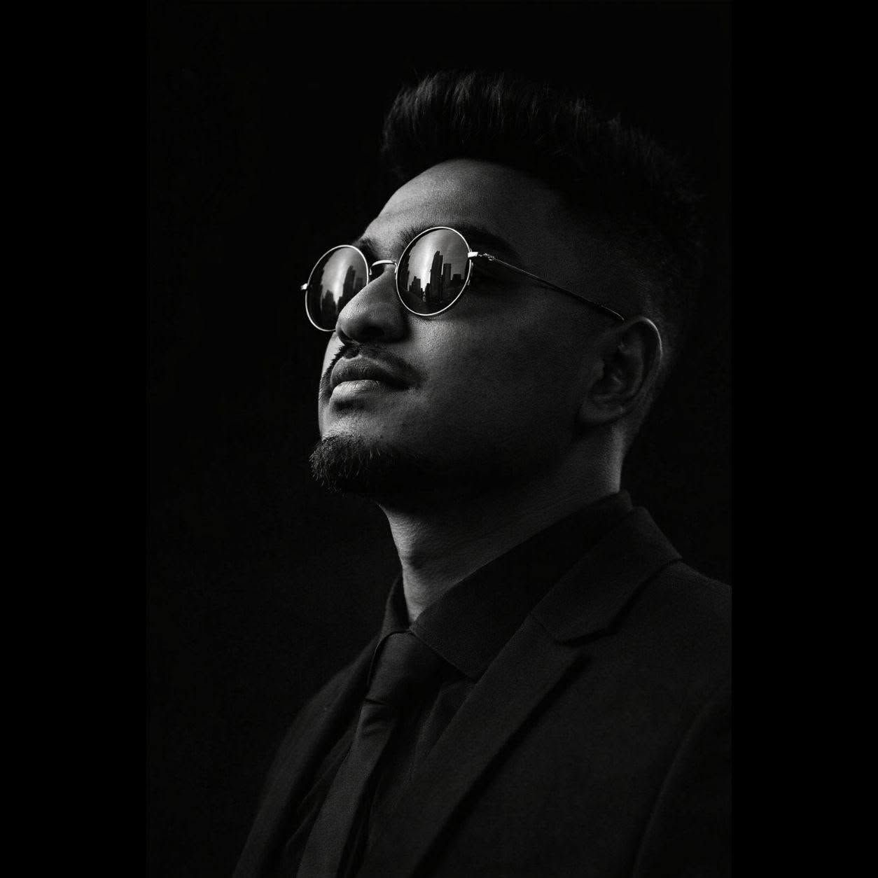 Black and white portrait of a stylish man wearing round sunglasses and a suit, gazing upward confidently.