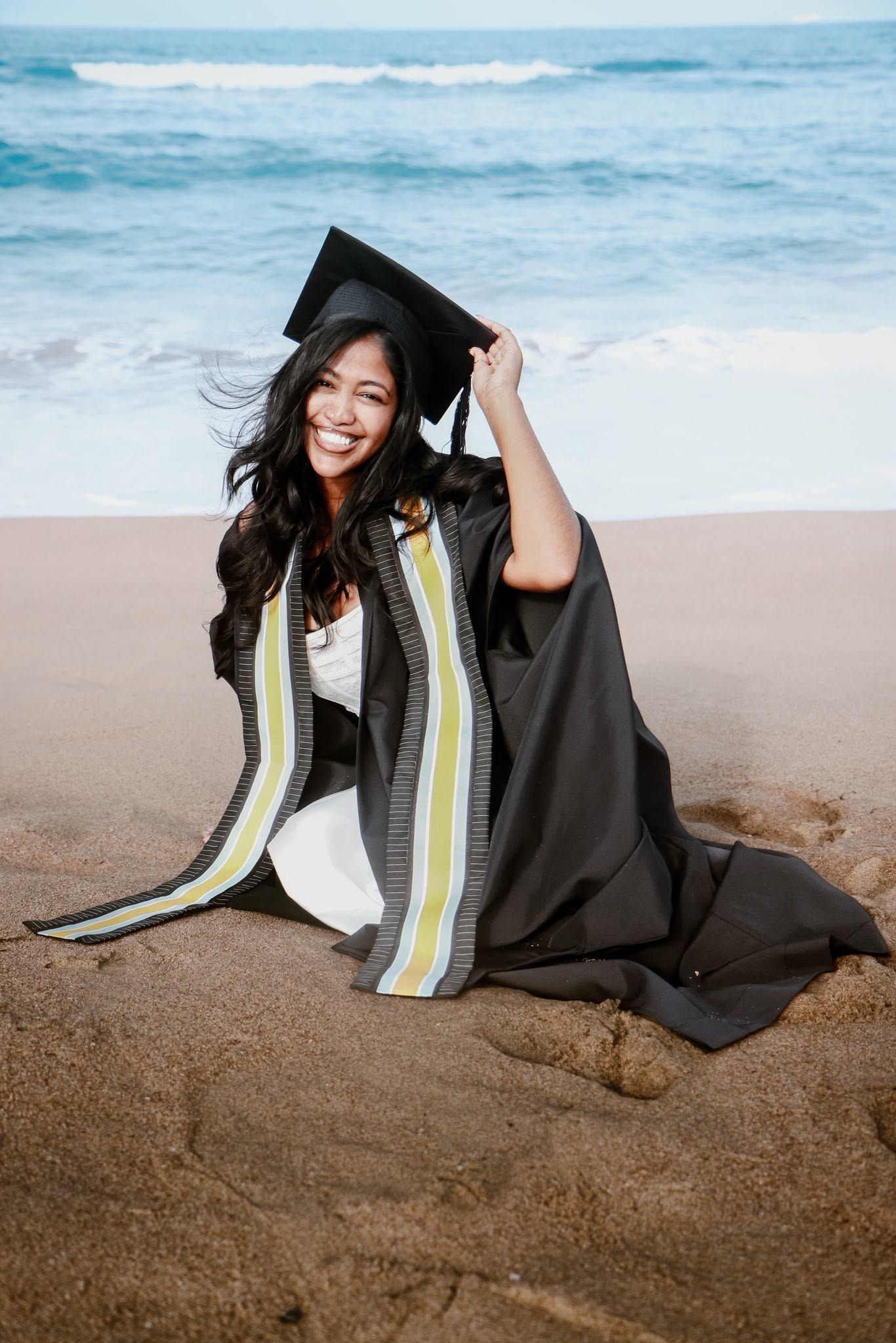 Smiling graduate in black cap and gown sitting on sandy beach with ocean waves in the background.