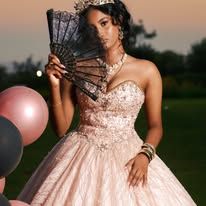 Elegant woman in rose gold ball gown holding black fan, wearing crown, posing outdoors at dusk.