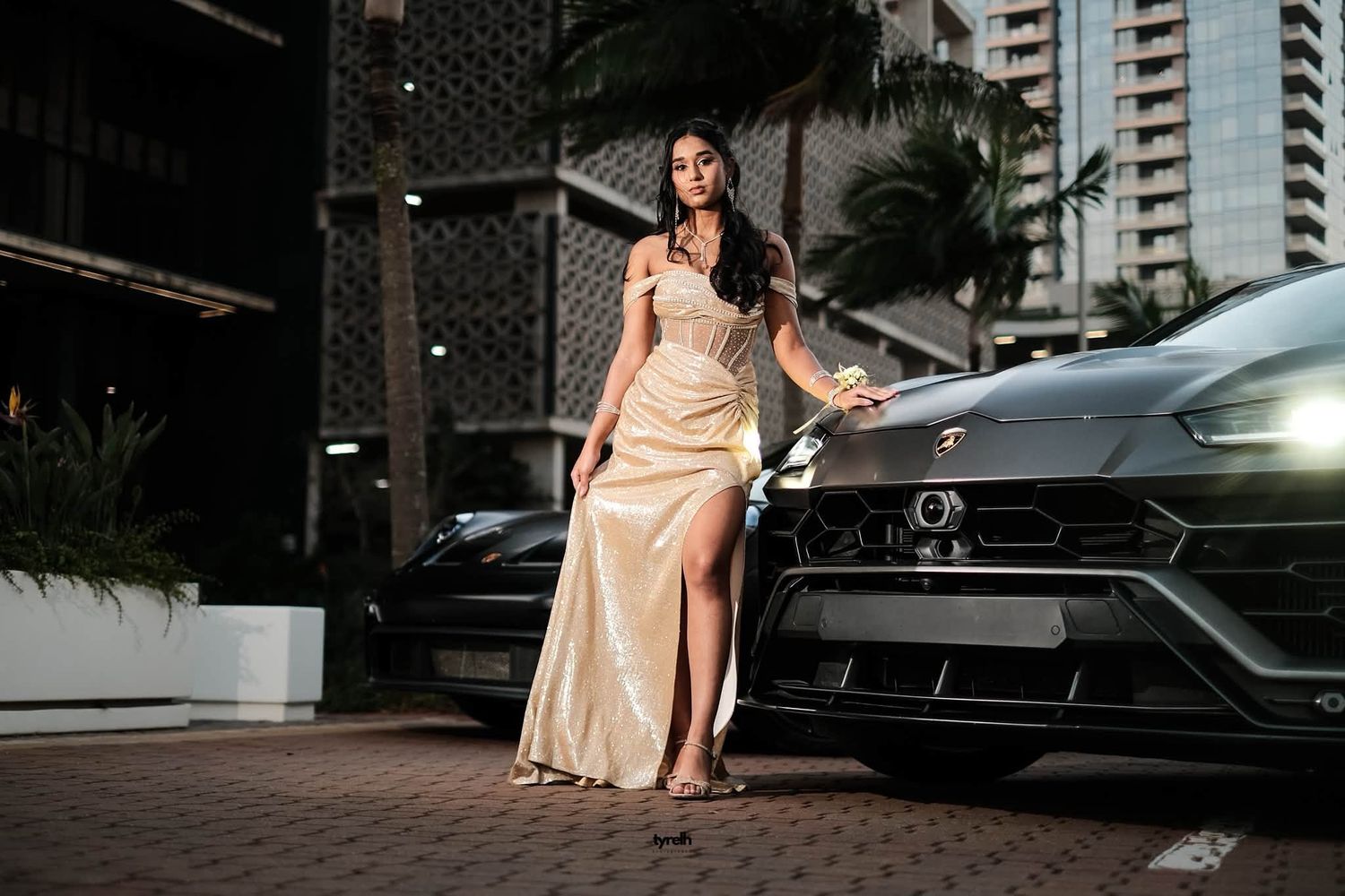 Elegant woman in gold slit gown posing beside a black Lamborghini Urus at night near luxury buildings.