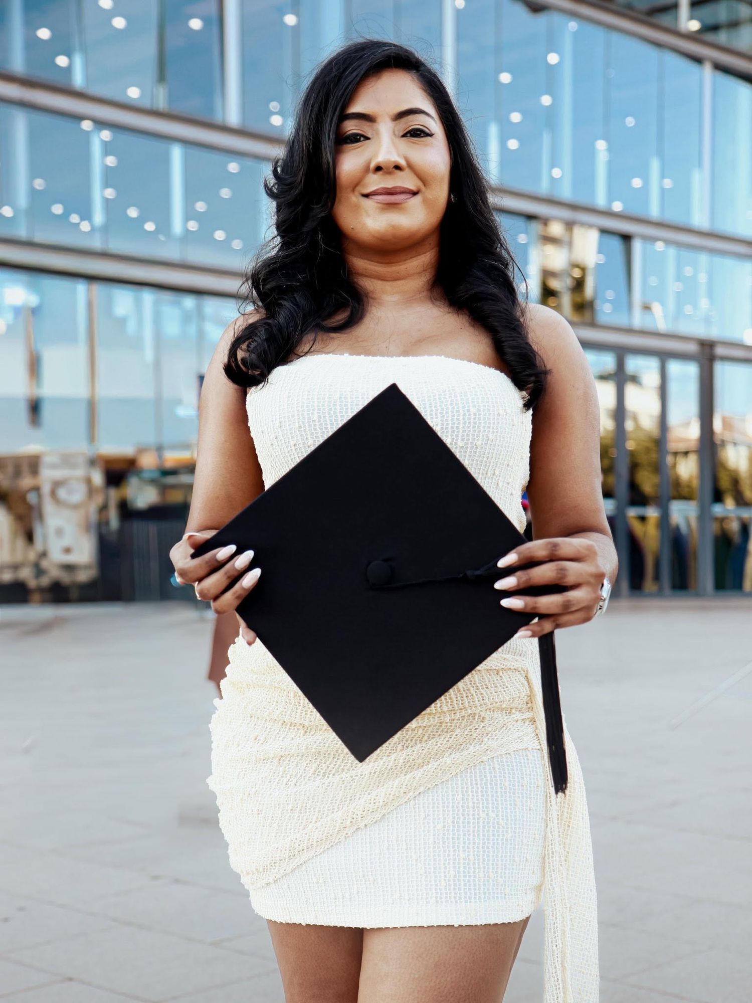 Young woman in white dress holding black graduation cap outdoors near modern glass building.