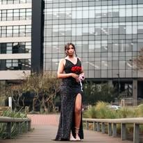 Woman in black sequin gown holding red roses poses on wooden walkway outside modern glass office building.