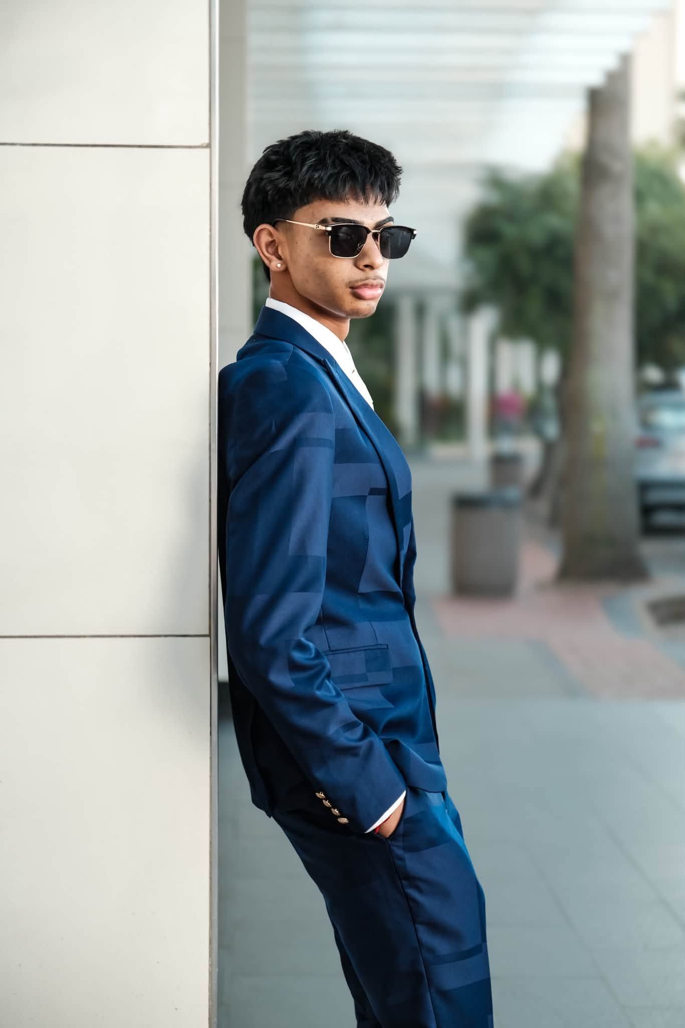 Young man in blue patterned suit and sunglasses leaning against wall on urban sidewalk.