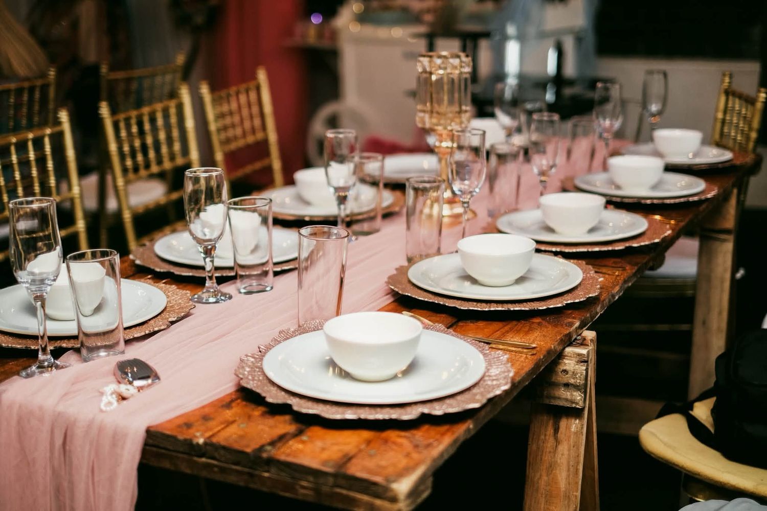 Elegant wedding reception table set with white china, pink runner, gold chairs, and crystal glassware on rustic wood table.