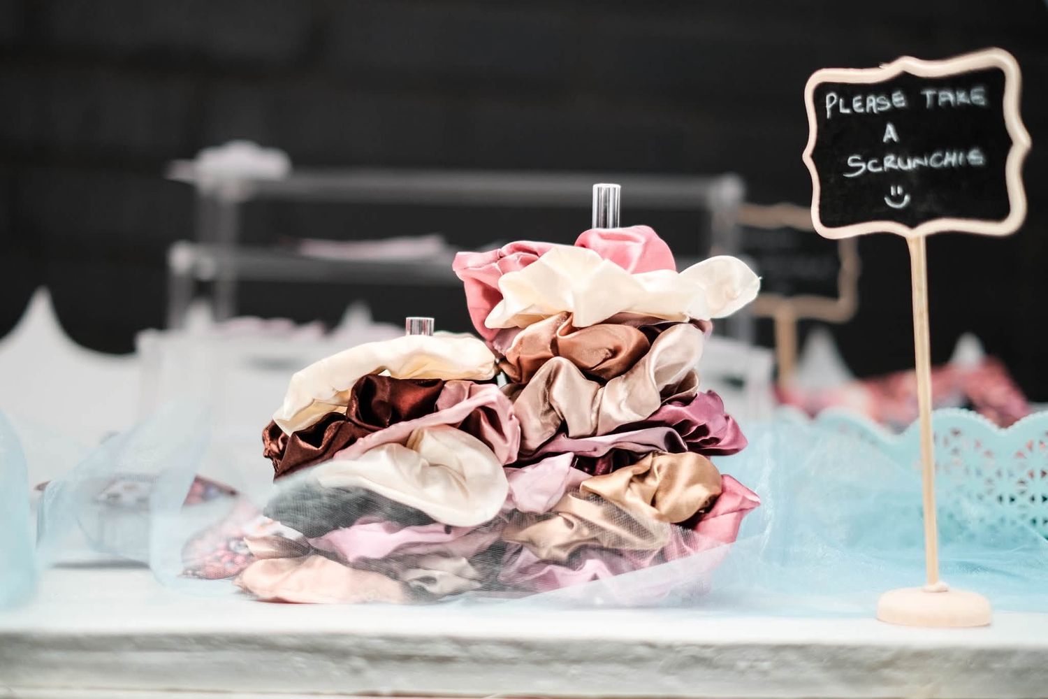 A pile of colorful hair scrunchies displayed on a table with a small chalkboard sign reading Please Take a Scrunchie.