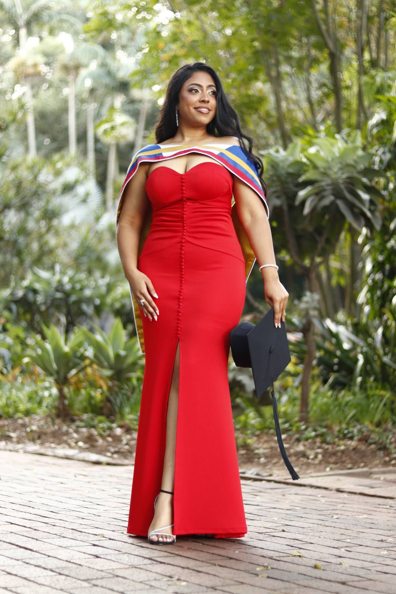 Woman in red off-shoulder gown holding graduation cap, posing outdoors among lush greenery on brick path.