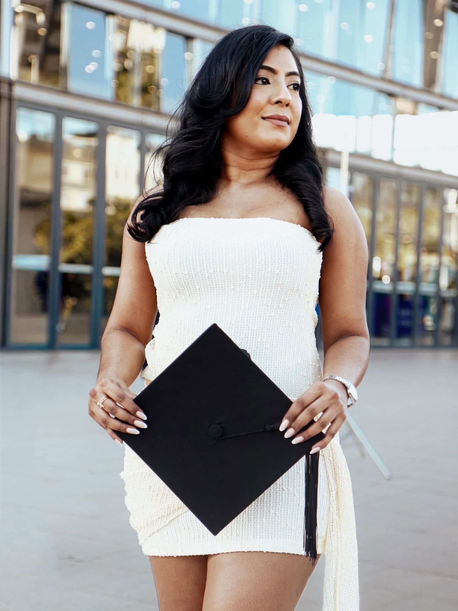 A young woman in a white strapless dress holds a black graduation cap outdoors, celebrating her academic achievement.