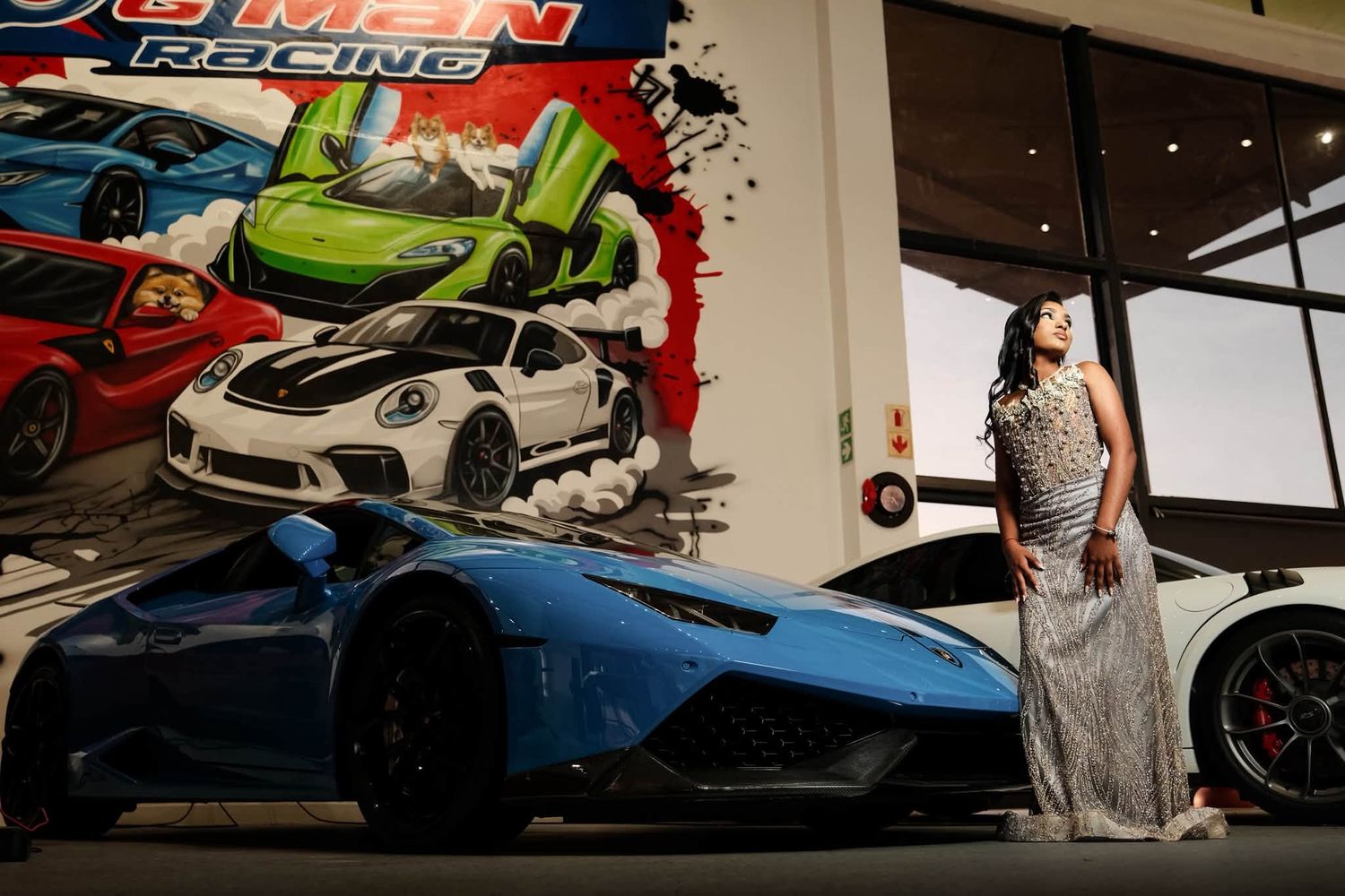 Woman in elegant gown poses beside a blue Lamborghini at an auto racing themed event with colorful car mural backdrop.