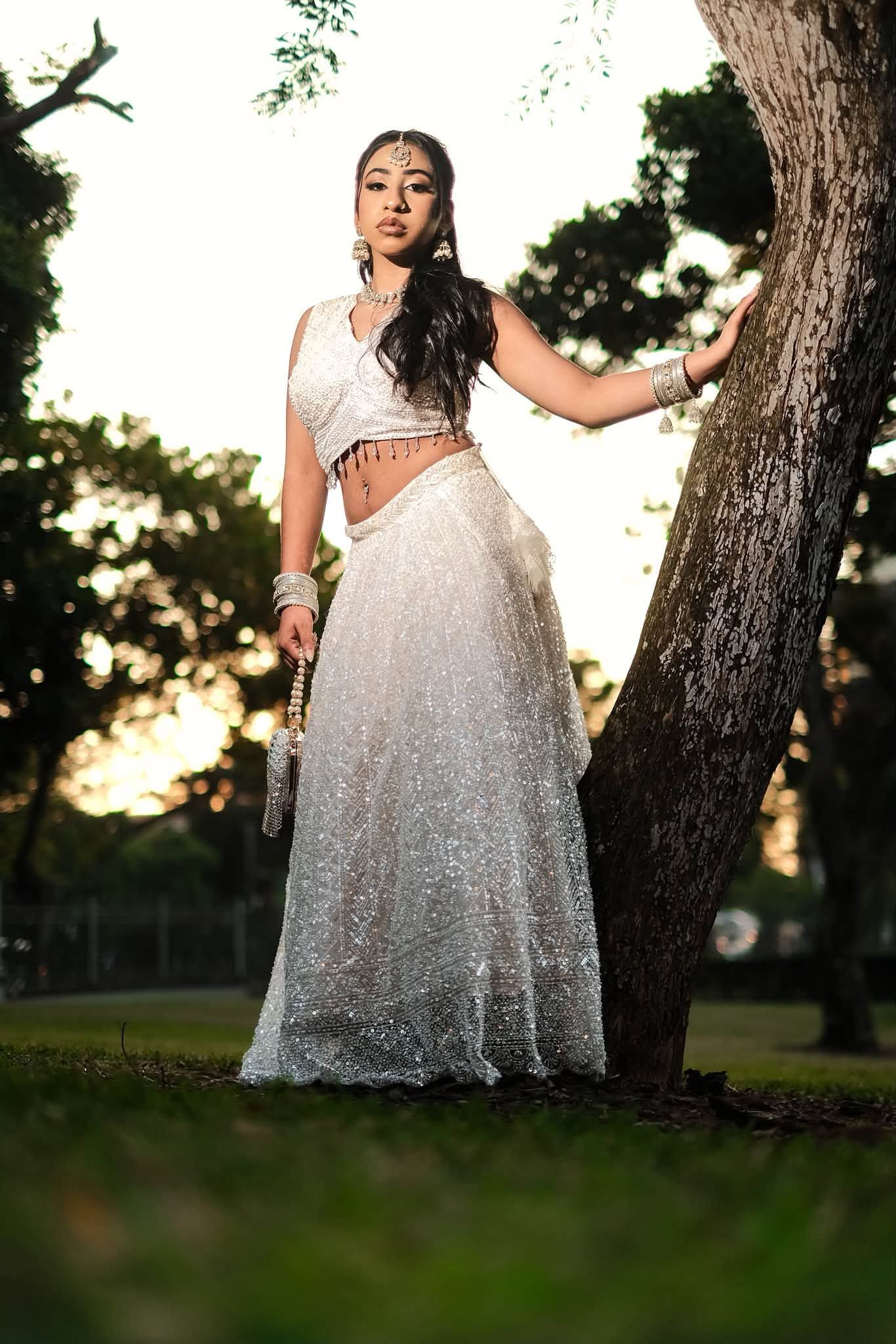 Young woman in white embellished Indian lehenga with jewelry poses gracefully by a tree in a park at sunset.