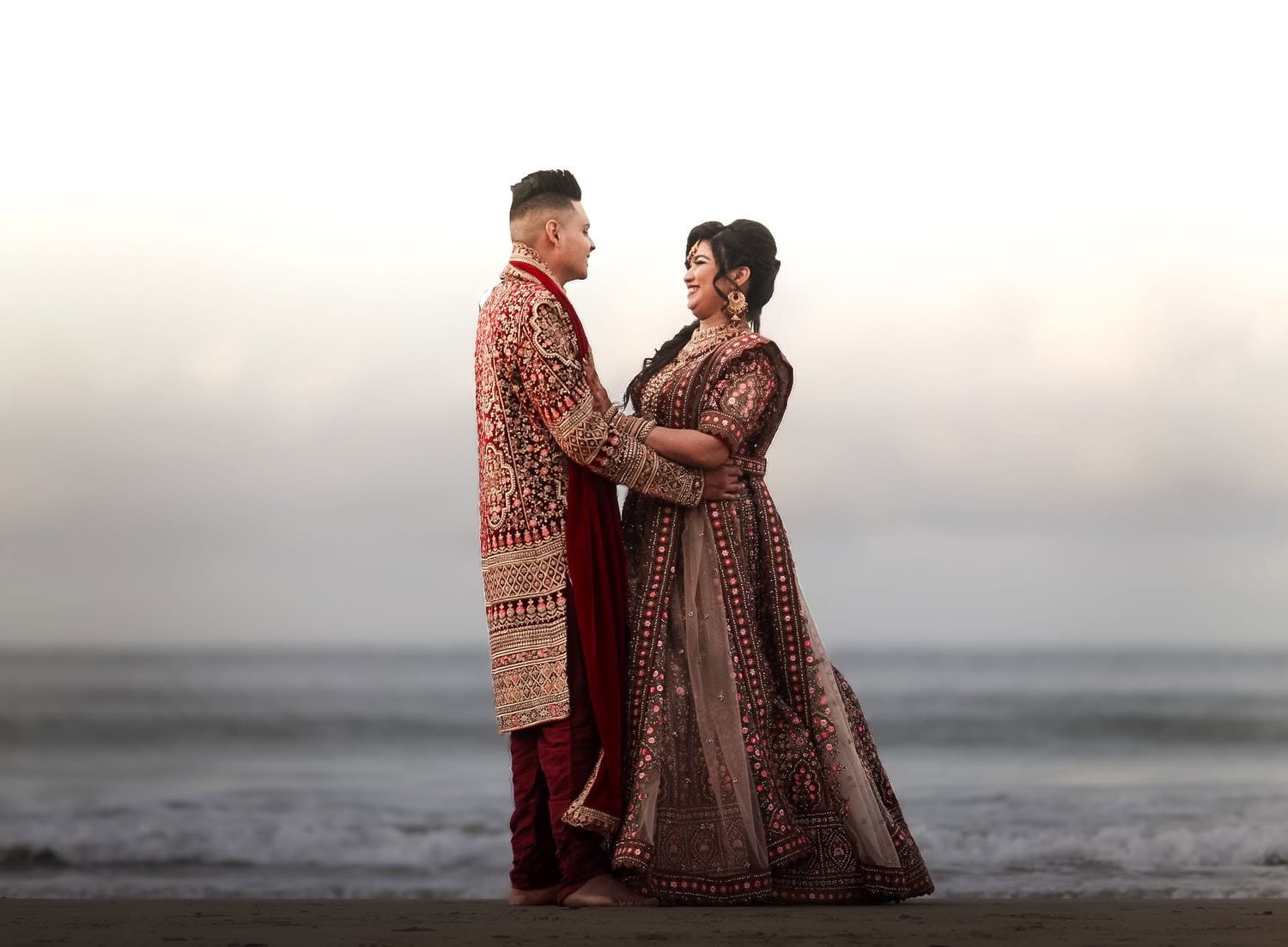 Indian couple in traditional red and maroon wedding attire posing on beach.