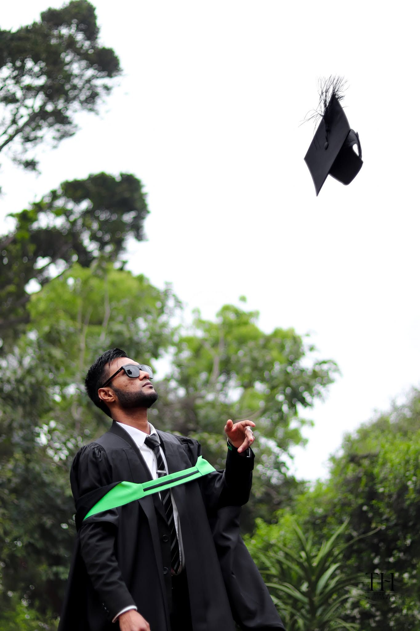 Graduate in black gown and sunglasses tossing mortarboard cap into the air outdoors among green trees.